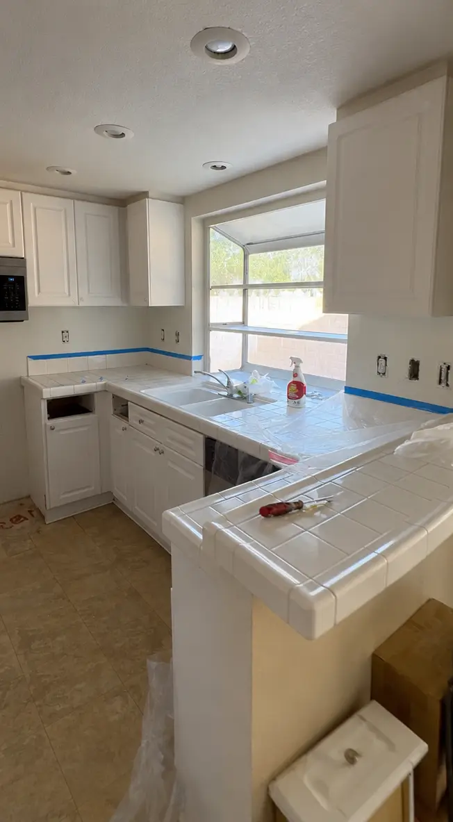 Before and after kitchen renovation by Eddie’s Ready Handyman & Home Improvement in Corona, CA, featuring freshly painted navy blue lower cabinets, white upper cabinets, a glossy gray subway tile backsplash, and updated window blinds while preserving the original countertops and kitchen layout. Convert to PDF Kitchen before cabinet painting and backsplash installation by Eddie’s Ready in Corona, CA, showing white cabinets and original tile countertops.