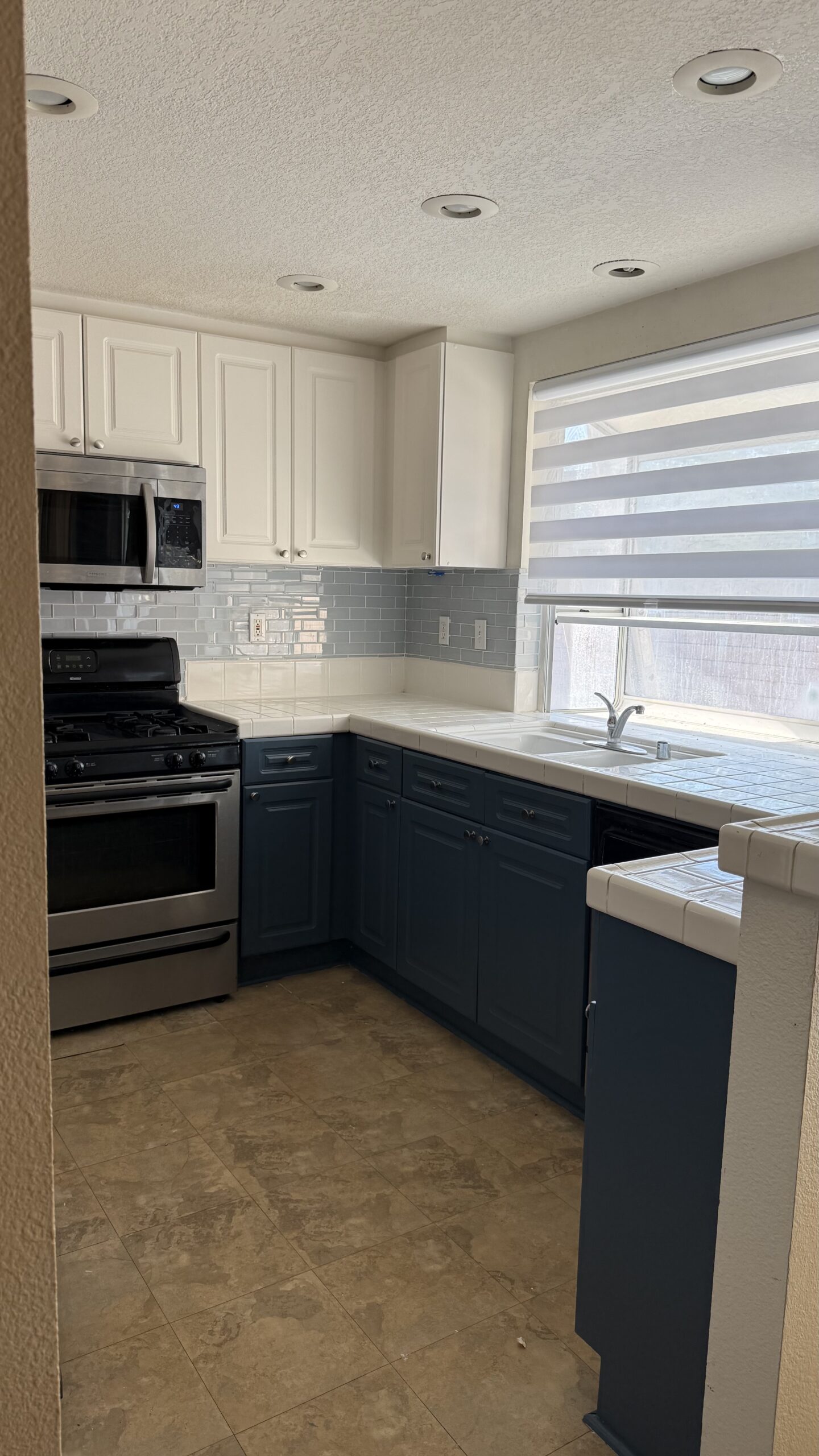 Kitchen after cabinet painting and gray subway tile backsplash installation in Corona, CA, featuring navy blue lower cabinets, white upper cabinets, and updated window blinds.